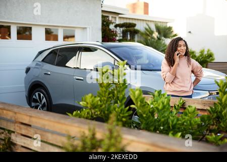 Giovane donna che parla con lo smartphone mentre si appoggia sull'auto nel cortile posteriore Foto Stock