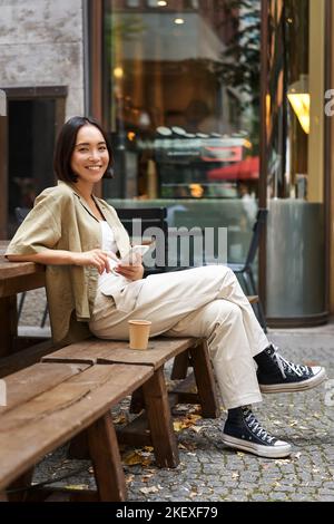 Ritratto di giovane donna coreana elegante siede in un bar, tiene smartphone, sorride, ama il caffè all'aperto Foto Stock