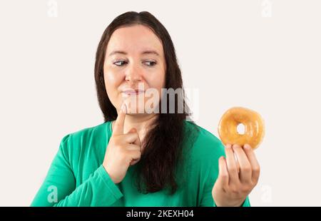 Donna ispanica di mezza età che mangia ciambella smaltata sopra isolato sfondo bianco faccia seria pensando alla domanda, idea molto confusa Foto Stock