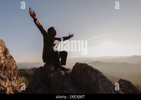 silhouette di uomo alzarsi mano in preghiera in cima alla montagna e tramonto cielo sfondo astratto. Concetto di libertà e avventura di viaggio. Foto Stock