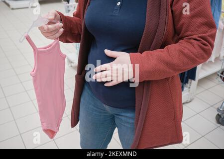 Donna incinta sceglie un vestito rosa - corpetto per un bambino in un negozio per acquistare Foto Stock