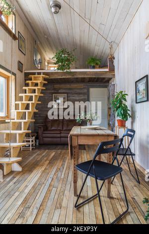 Tavolo da pranzo in legno con sedie pieghevoli e scale retrattili in legno di pino che conducono alla camera da letto soppalcata e all'area soggiorno con divano in pelle marrone. Foto Stock