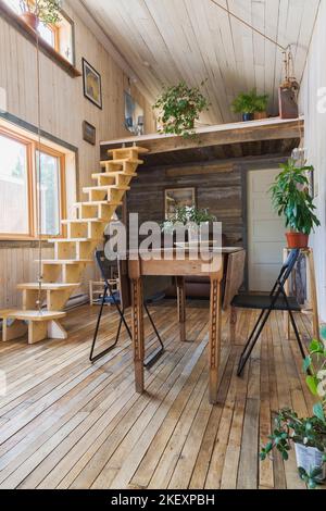 Tavolo da pranzo in legno con sedie pieghevoli e scale retrattili in legno di pino che conducono alla camera da letto soppalcata e all'area soggiorno con divano in pelle marrone. Foto Stock