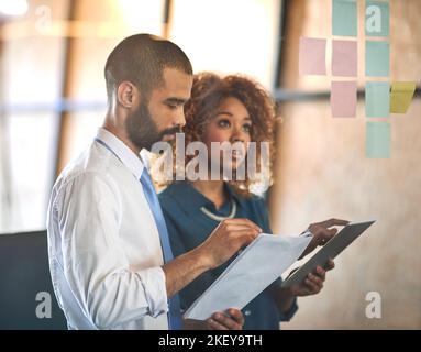 Imprenditori che fanno quello che fanno meglio. Due giovani professionisti che fanno il brainstorming con note appiccicose su una parete di vetro in un ufficio. Foto Stock