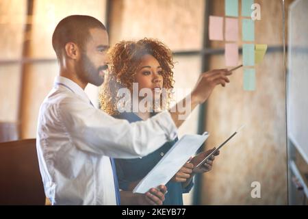 Imprenditori che fanno quello che fanno meglio. Due giovani professionisti che fanno il brainstorming con note appiccicose su una parete di vetro in un ufficio. Foto Stock