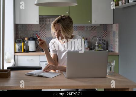 La donna disoccupata legge la lista di scrittura del taccuino dei contatti, cercando i posti vacanti sul laptop sul tavolo Foto Stock