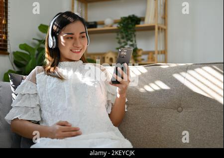 Attraente donna asiatica incinta indossando cuffie, ascoltando musica o guardando video neonato cura del bambino su Internet mentre si siede sul divano in lui Foto Stock