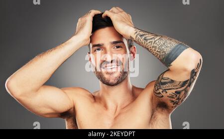 Viso, tatuaggio e bellezza con un modello uomo in studio su sfondo grigio per il benessere con fiducia. Ritratto, felice e sorridente con un bel giovane Foto Stock