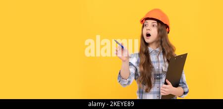 Bambino di rinnovamento, scioccata ragazza teen in casco e camicia a scacchi facendo appunti sulla clipboard, lavorando. Bambino con disegno orizzontale del poster dell'elmetto Foto Stock