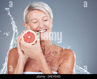 Cura della pelle, acqua e ritratto di vecchia donna con pompelmo in background grigio studio per il benessere. Bellezza, spruzzi d'acqua e donna anziana con frutta Foto Stock