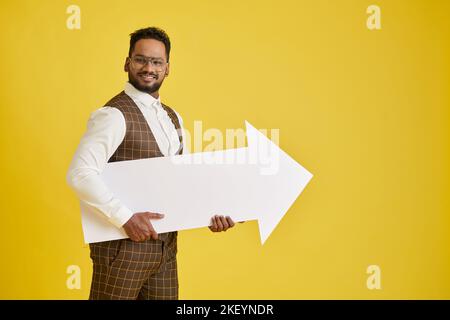 Uomo d'affari eccitato che tiene la freccia bianca enorme del foglio, isolato sul giallo Foto Stock