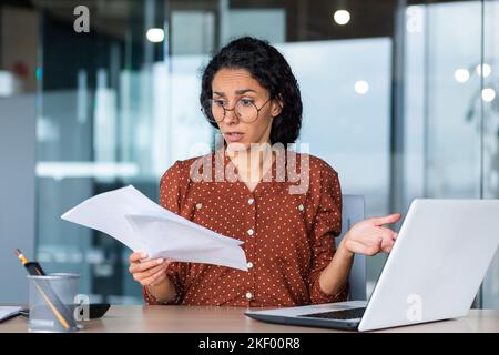 Donna d'affari frustrata e triste che tiene il rapporto finanziario nelle mani, donna ispanica triste che lavora in ufficio usando il laptop, contabile finanziere femminile infelice con i risultati di lavoro. Foto Stock