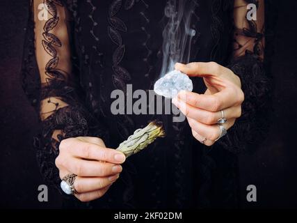 Vista ravvicinata della donna in abito di pizzo nero, gemma di cristallo celeste di pulizia macchiando il fascio di salvia bianco. Rimuovere l'energia negativa. Foto Stock