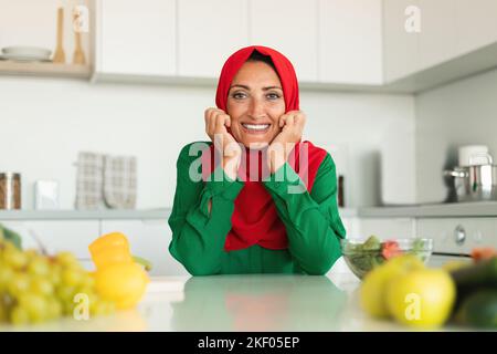 Ritratto di felice casalinga musulmana di mezza età in posa in cucina, seduta a tavola e sorridente alla macchina fotografica, spazio libero Foto Stock
