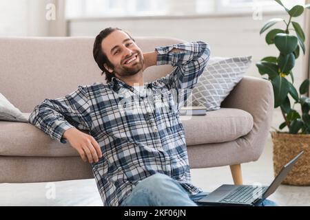 Felice uomo europeo millenario in abiti domestici con stoppia siede sul pavimento, lavorando sul computer in soggiorno Foto Stock