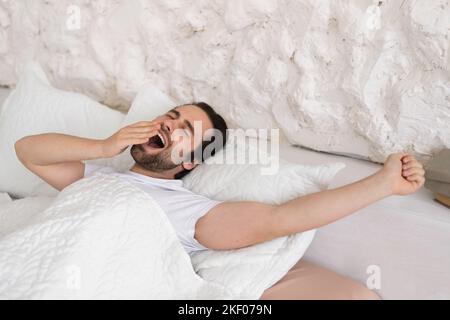 Allegro uomo europeo millenario con stoppia si sveglia, si sbadigna sul letto bianco, godere di un ambiente accogliente, confortevole e pigro Foto Stock