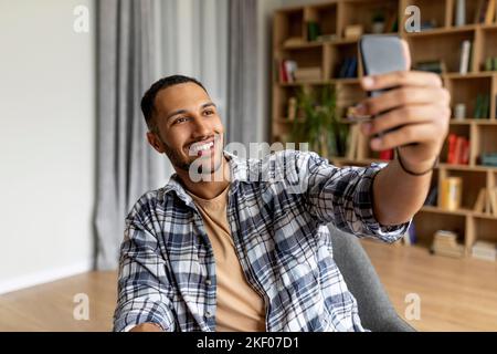 Buon giovane arabo che videochiama via smartphone o fa selfie, seduto in poltrona a casa, copy space Foto Stock