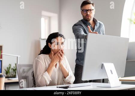 Il giovane uomo arrabbiato europeo, capo, spaventa la triste manager signora sul posto di lavoro con il computer Foto Stock