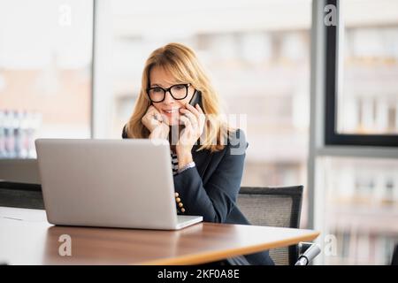 Donna d'affari di mezza età che utilizza un computer portatile ed effettua una chiamata mentre si siede alla scrivania in ufficio. Donna d'affari executive che indossa occhiali e blazer. Foto Stock