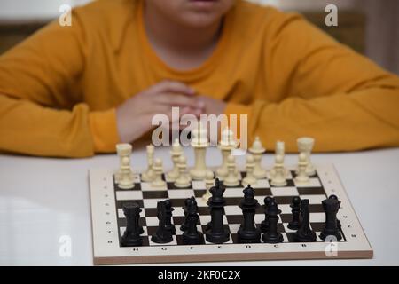 siede a un tavolo e gioca a scacchi. Il bambino si è concentrato sul gioco e pensa dove fare la sua prossima mossa. Sviluppo precoce, giochi educativi domestici Foto Stock