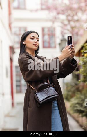 donna brunette con auricolare wireless e tracolla che prende selfie a praga, immagine stock Foto Stock