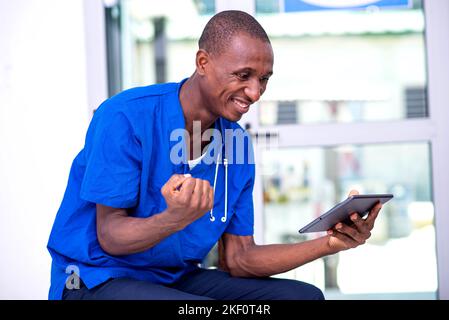 giovane medico maschile seduto sulla sedia utilizzando un tablet digitale facendo un gesto vincente mentre sorrida. Foto Stock