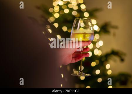 Mano che tiene un bicchiere di vino, le luci di natale sfocate sull'albero di natale sullo sfondo. Pino decorato con ghirlanda splendente, persona tenuta Foto Stock