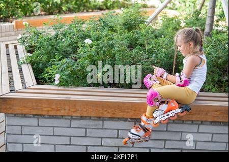 Una bambina caucasica in pattini a rotelle siede su una panca. Foto Stock