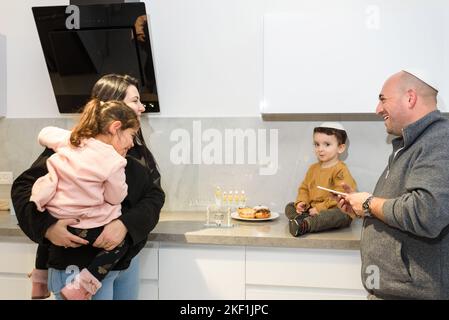 Genitori felici con i bambini che illuminano le candele sulla menorah tradizionale alla cucina, celebrando Hanukkah nel paese. Il Padre recita le benedizioni di Hanukkah e illumina le candele sulla menorah. Foto Stock