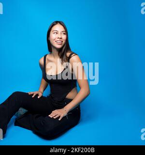 Bella multirazziale Teen femmina seduto Croce in gamba sul pavimento guardando a destra Copy Space su sfondo isolato blu Foto Stock