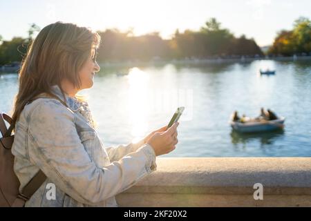 Donna che guarda il suo telefono cellulare accanto a un lago nel parco e con luce solare che illumina la scena da dietro. Foto Stock