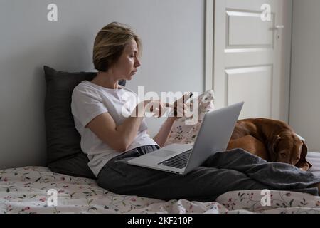 Donna di mezza età si siede sul letto con laptop che chiama il potenziale datore di lavoro sul telefono vicino al cane Foto Stock