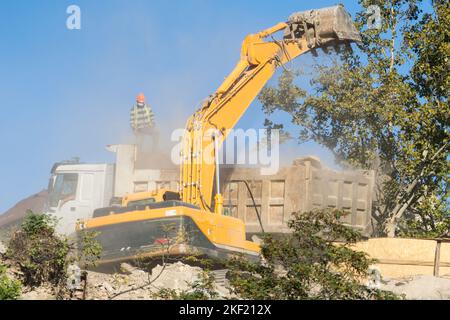 Demolizione dell'escavatore di vecchi alloggiamenti fatiscenti e caricamento dei rifiuti di costruzione nel dumper posteriore Foto Stock