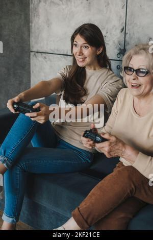 Figlia matura allegra con la madre anziana che tiene controller per videogiochi, che si diverte con i giochi virtuali e che ride insieme. Concetto di Home Entertainment Foto Stock