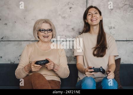 Famiglia felice: Figlia e sua madre caucasica che gioca a giochi da console nel soggiorno seduto sul divano. Stile di vita delle persone. Famiglia felice. Attività Foto Stock