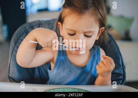 Allegra bambina che mangia da sola con il cucchiaio, mentre si siede su una seggiolone. Ritratto di ragazzo felice. Mangiare sano. Foto Stock