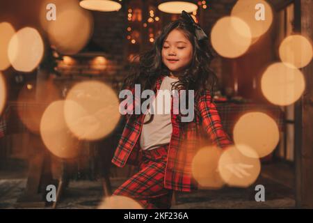 Attiva ragazza bambino giappone con elegante modello di capelli brunette lunga posa a casa in bokeh ghirlands noel. Asian piccolo fashionista in rosso plaid pajama danza Foto Stock