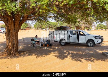 Sesriem Campsite, Sossusvlei, Namibia - 01 ottobre 2018: Una ragazza seduta sotto un albero di acacia nel campeggio. Auto con attrezzature parcheggiate nelle vicinanze. Foto Stock