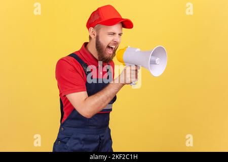 Vista laterale di un operaio arrabbiato che tiene il megafono vicino alla bocca a voce alta, urlando, protestando, indossando uniforme blu e cappuccio rosso. Studio al coperto isolato su sfondo giallo. Foto Stock