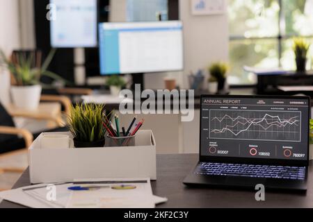 Società di analisi finanziaria con postazioni di lavoro vuote prima dell'arrivo del personale la mattina presto. Computer portatile sulla scrivania con rapporto annuale sulle vendite, schermi elettronici in background. Foto Stock