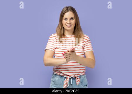 Ritratto di una donna bionda positiva sorridente che indossa una T-shirt a righe in piedi con le mani, stringendo le mani, applaudendo. Studio al coperto isolato su sfondo viola. Foto Stock