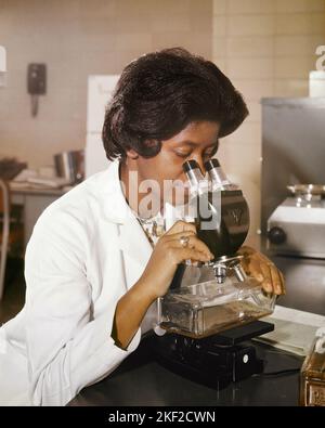 1960S AFRO-AMERICANA DONNA TECNICO DI RICERCA AL MICROSCOPIO SCANSIONE PER LE CELLULE TUMORALI IN UNA COLTURA - KM1096 HAR001 HARS OCCUPAZIONI SCANSIONE CELLULE DI COLTURA MID-ADULT DONNA MID-ADULT HAR001 LABORATORI ANTICHI AFRO-AMERICANI Foto Stock