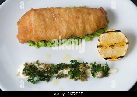 Pesce e patatine servite su una crema di piselli verdi e un'insalata di maionese al cappero. Foto Stock