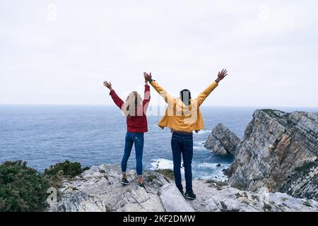 ragazzo latino e ragazza caucasica in giacca rossa e jeans gialli in piedi indietro tenendo le mani con le braccia distese dalle grandi rocce, cabo de Foto Stock