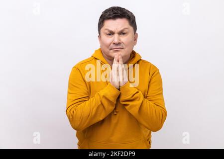 Prego, sto pregando. Ritratto di bruna rovesciato uomo che tiene le braccia in preghiera, chiedendo aiuto o perdono con gli occhi imploranti, indossando felpa con cappuccio stile urbano. Studio in interni isolato su sfondo bianco. Foto Stock