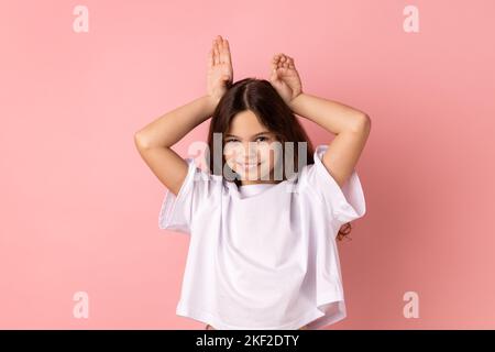 Ritratto di bambina positiva che indossa una T-shirt bianca che fa divertenti orecchie da coniglietto con le mani sulla testa, comportamento infantile, allegro umore ottimista. Studio in interni isolato su sfondo rosa. Foto Stock