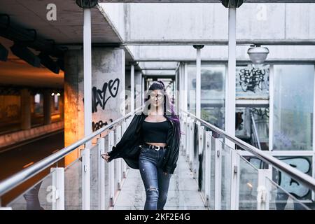 latina ragazza con occhiali lunghi in pelliccia brunetta giacca strappato jeans trafitto labbro camminare sul passaggio di vetro Foto Stock