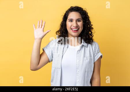 Ritratto di amichevole positiva bella donna con i capelli ondulati scuri sorride dentifricamente, solleva palma saluta amico, dicendo ciao o Arrivederci. Studio al coperto isolato su sfondo giallo. Foto Stock