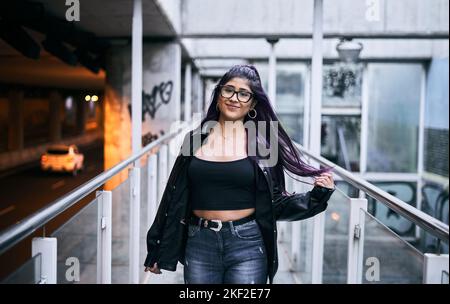 latina ragazza con occhiali bruna pelliccia giacca lunghi capelli tondi orecchini labbro piercing sorridente alla macchina fotografica che cammina lungo il passaggio urbano Foto Stock