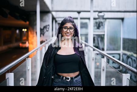 latina ragazza con occhiali in pelliccia brunetta collana giacca sul collo orecchini tondi labbro piercing sorridente alla fotocamera che cammina lungo il passaggio urbano Foto Stock